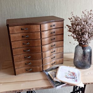 May include: A wooden chest of drawers with a curved front and multiple drawers. The chest is brown with dark metal handles. A vase of dried flowers and a cross-stitch project are next to the chest.