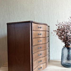 May include: A wooden jewelry box with a curved front and multiple drawers. The box has a dark brown finish on the sides and a lighter wood finish on the drawers. Each drawer has a small, ornate handle.