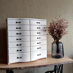 May include: A white wooden chest of drawers with eight drawers and black handles. The chest sits on a wooden table with a decorative vase of dried flowers. The background is a textured, neutral-toned wall.