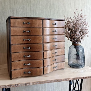 May include: A wooden jewelry chest with twelve drawers, each with a curved front and a small, ornate handle. The chest is a warm brown color and sits on a wooden table. A decorative vase with dried flowers is to the right.