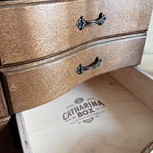 May include: Close-up of a wooden jewelry box with three drawers. The drawers are a warm brown color with decorative black metal handles. The bottom drawer is open, revealing the text "CATHARINA BOX" inside.