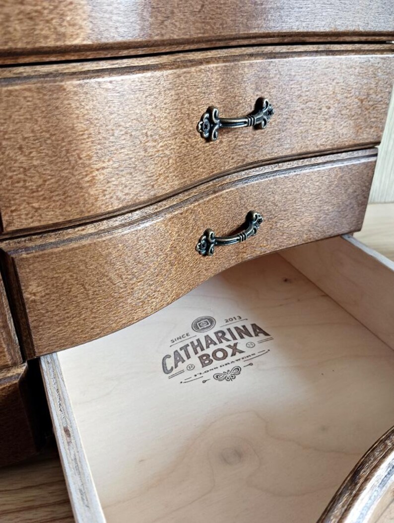 May include: Close-up of a wooden jewelry box with three drawers. The drawers are a warm brown color with decorative black metal handles. The bottom drawer is open, revealing the text "CATHARINA BOX" inside.