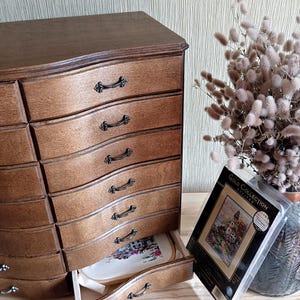 May include: A wooden chest of drawers with eight curved drawers, each with a black handle. One drawer is open, revealing a cross-stitch project in a wooden hoop. A Gold Collection cross-stitch kit is visible.
