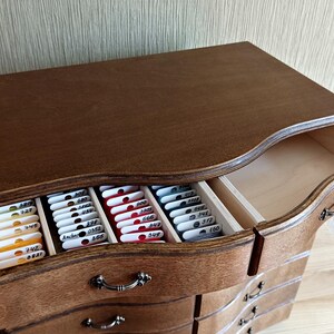May include: A wooden chest of drawers with a dark brown finish. The top drawer is open, revealing organized compartments filled with colorful thread spools. The chest has a curved design and ornate metal handles, ideal for craft storage.
