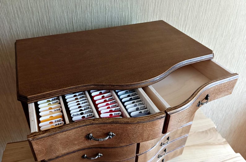 May include: A wooden chest of drawers with a dark brown finish. The top drawer is open, revealing organized compartments filled with colorful thread spools. The chest has a curved design and ornate metal handles, ideal for craft storage.