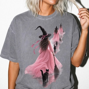 May include: A charcoal grey short-sleeve t-shirt featuring a graphic print of a figure in a pink dress and black witch hat holding a broom, accompanied by black cats. The design shows multiple figures walking away, creating a repeating visual effect.