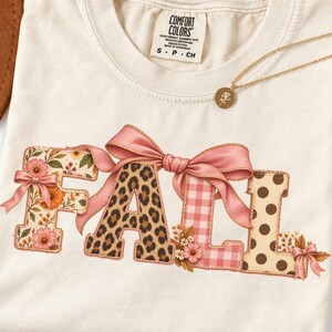 May include: A cream-colored t-shirt featuring the word FALL in large letters. The F has a floral print, the A is leopard print, the L is pink gingham, and the L has brown polka dots. A large pink ribbon bow sits atop the letters.