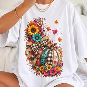 May include: A white t-shirt featuring a central graphic of a pumpkin decorated with leopard print, black and white buffalo plaid, and floral patterns. The pumpkin is accented with a large buffalo plaid bow and surrounded by sunflowers and autumn leaves.
