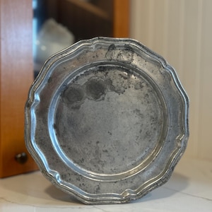May include: A silver-toned pewter plate with a scalloped edge. The plate has a slightly worn, antique look.