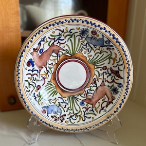 May include: A round ceramic plate with a colorful floral design featuring two deer, birds, and a central white circle. The plate has a brown rim with a blue and yellow border.