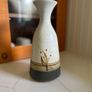 May include: A handmade ceramic vase with a speckled white body, a brown and tan band, and a dark gray base. The vase features a decorative leaf design and a textured, flared rim. This decorative vase is perfect for displaying flowers or as a standalone art piece.