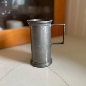 May include: A pewter-colored metal mug with a rectangular handle. The mug has a cylindrical body and a slightly flared base. The handle is attached to the side of the mug. The mug is sitting on a white surface.
