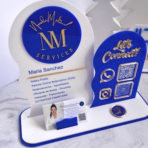 May include: A white and blue acrylic business display sign featuring the text Nanda Multi Services, NM, and Maria Sanchez. It includes a business card holder, gold-colored social media icons, QR codes, and a circular NFC tag on a rectangular base.