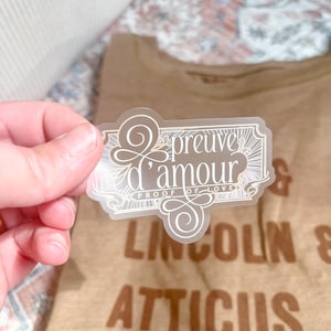 May include: A clear sticker with the text "preuve d'amour" and "proof of love" in a decorative frame. The sticker is held in front of a brown t-shirt with the words "Lincoln & Atticus" printed on it.