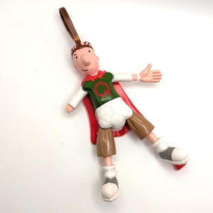 May include: A plastic keychain figure of a cartoon character with brown hair, wearing a green shirt with a red letter Q, a red cape, white shorts, and brown shorts over them. The figure has light skin, white sleeves, and gray and white shoes.