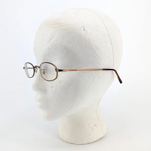 May include: Oval-shaped eyeglasses with a brown metal frame and temples. The Calvin Klein logo is visible on the temple. The glasses are displayed on a white foam head.