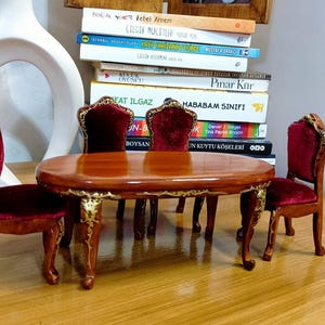 Pode incluir: Um conjunto de jantar em miniatura com uma mesa de madeira castanho e quatro cadeiras. As cadeiras têm assentos de veludo vermelho e costas ornamentadas. A mesa tem uma borda esculpida e detalhes dourados.