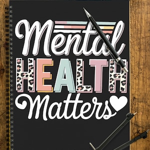 Könnte beinhalten: Schwarzes Spiralnotizbuch mit den Worten "Mental Health Matters" in Weiß, Rosa und Blau. Ein schwarzer Stift und ein Zirkel liegen auf dem Notizbuch. Das Notizbuch liegt auf einer Holzoberfläche.