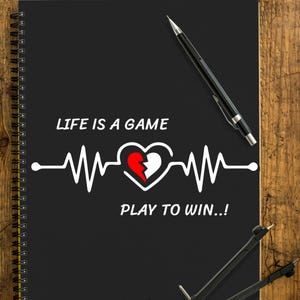 Könnte beinhalten: Schwarzes Spiralnotizbuch mit dem Text "LIFE IS A GAME PLAY TO WIN..!" in Weiß. Das Design umfasst eine Herzschlagsymbol-Linie und ein Herz mit einem roten, gebrochenen Abschnitt. Stifte, ein Zirkel und ein Lineal liegen auf einer Holzoberfläche.
