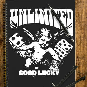 Könnte beinhalten: Schwarzes Spiralnotizbuch mit dem weißen Wort "UNLIMITED", über einer Grafik eines Engels, Würfeln und den Worten "GOOD LUCK". Ein Bleistift und ein Zirkel liegen auf einer Holzoberfläche.