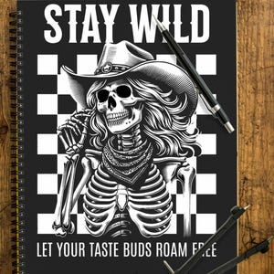 Könnte beinhalten: Schwarzes Spiralnotizbuch mit einer Skelett-Cowboy-Grafik. Das Bild zeigt den Text "STAY WILD" und "LET YOUR TASTE BUDS ROAM FREE". Das Notizbuch liegt auf einer Holzoberfläche mit Stiften und einem Zirkel.
