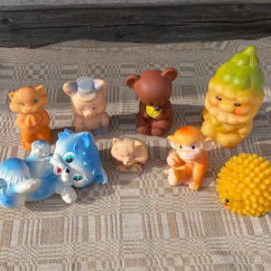 Puede incluir: Colección de juguetes de goma vintage en varias formas y colores. Incluye un gato azul, un zorro naranja, un oso marrón, un mono amarillo y una bola espinosa amarilla. Los juguetes están colocados sobre una superficie estampada.