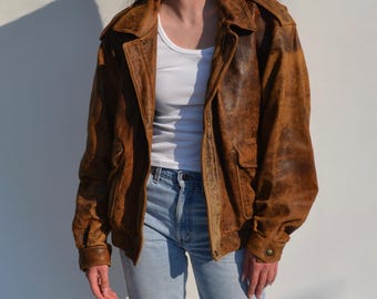 Op de afbeelding: Een bruine leren jas met een distressed afwerking. De jas heeft een kraag, twee voorzakken en manchetten met knopen. De jas wordt gedragen over een witte tanktop en een blauwe jeans.