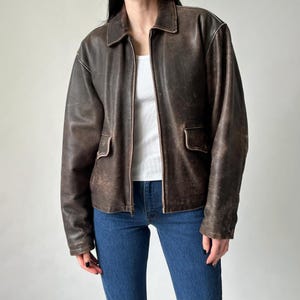 Op de afbeelding: Bruine leren jas met een distressed afwerking. De jas heeft een kraag, twee voorzakken en een ritssluiting. Het model draagt een witte top en een blauwe jeans.