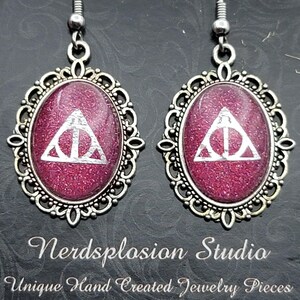 May include: Silver-coloured earrings with oval, glittery magenta centres. Each centre displays a white Deathly Hallows symbol within a decorative filigree frame. The text "Nerdsplosion Studio" and "Unique Hand Created Jewelry Pieces" are below the earrings.