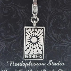 May include: Silver-toned charm featuring the Sun tarot card design. The rectangular pendant displays a sunburst with the words "THE SUN" at the bottom. It is attached to a clasp with a black cord. The text "Nerdsplosion Studio" and "Unique Hand Created Jewelry Pieces" are also visible.
