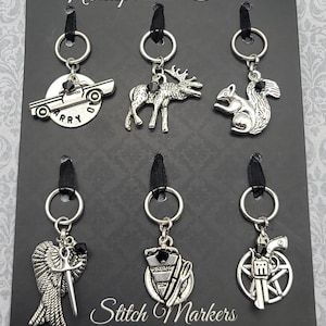 May include: A set of six silver-toned stitch markers with various charms, including a car, moose, squirrel, angel with a sword, a pie with utensils, and a gun with a pentagram. The markers hang from black ribbons and silver rings. The card reads "Nerdsplosion Studio" and "Stitch Markers".