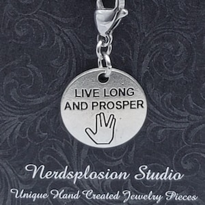 Puede incluir: Un dije plateado con las palabras "LIVE LONG AND PROSPER" y un gesto con la mano. El dije está sujeto a un cordón negro con un cierre plateado. El fondo es una superficie oscura y estampada.