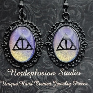 May include: A pair of black framed earrings with an oval design. The design features a black Deathly Hallows symbol on a purple and gold background. The earrings are from Nerdsplosion Studio, unique hand-created jewelry pieces.