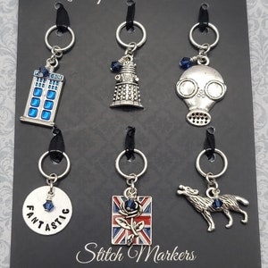 May include: A collection of silver-toned stitch markers with black ribbons and silver rings. The markers feature various charms, including a blue police box, a Dalek, a gas mask, a disc with the word "FANTASTIC", a Union Jack with a rose, and a wolf.