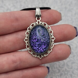 May include: Oval pendant with a silver-colored filigree frame and a black and purple floral design. The pendant hangs from a black cord. The design features a swirling pattern in shades of purple against a dark background.