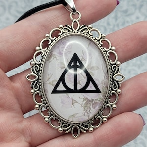 May include: Oval pendant with a silver-colored filigree frame. The pendant features a black Deathly Hallows symbol on a floral background. It hangs from a black cord.