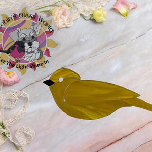 May include: A stained glass bird ornament in a solid mustard yellow color with a small black beak and a white eye detail. Beside the bird is a circular sticker featuring a dog illustration and the text Robin's Glass World Let The Light Shine In.