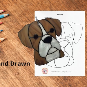 May include: A stained glass suncatcher shaped like a boxer dog head with brown and white glass pieces, displayed next to a printed paper pattern labeled BOXER and Hand Drawn, featuring black outlines and the text 2016 Robin C. Hary All Rights Reserved.