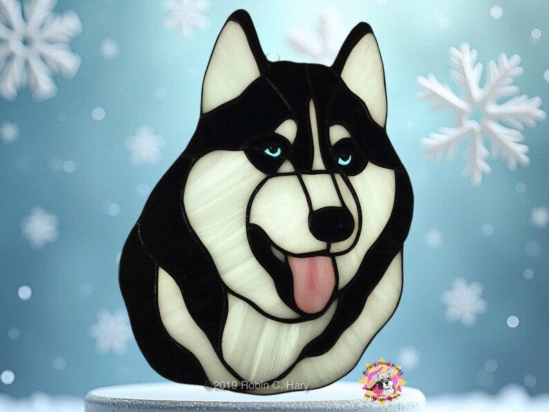 May include: Stained glass art of a black and white husky dog with blue eyes. The dog is looking to the right and has its tongue sticking out. The background is a light blue color with snowflakes.
