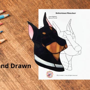 Puede incluir: Una pieza de vitral de un Doberman Pinscher con segmentos de vidrio negro, canela y rosa, junto a un patrón de papel blanco etiquetado Doberman Pinscher, Level: Beginner, Size: w: 12,7 cm x h: 20,3 cm. El texto Hand Drawn es visible a la izquierda.