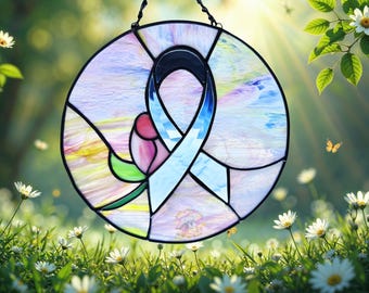 May include: A circular stained glass suncatcher featuring a light blue awareness ribbon and a pink rose with a green stem. The design is composed of various glass segments in shades of pastel pink, blue, and white, all held together by black metal cames.