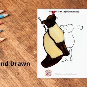 Op de afbeelding: Een glas-in-loodpatroon met een kat met een kristallen vlinder. De afbeelding staat op een wit vel papier met de tekst "Siamese with Crystal Butterfly". Kleurpotloden liggen op een houten oppervlak. De woorden "Hand Drawn" zijn ook zichtbaar.