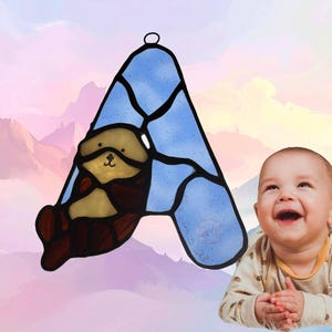 Puede incluir: Un atrapasoles de vidrieras con forma de letra A, con un fondo de cristal texturizado azul claro y una ilustraci&oacute;n de una nutria marina marr&oacute;n y beige. La pieza est&aacute; delineada con soldadura de metal negro e incluye un lazo para colgar.