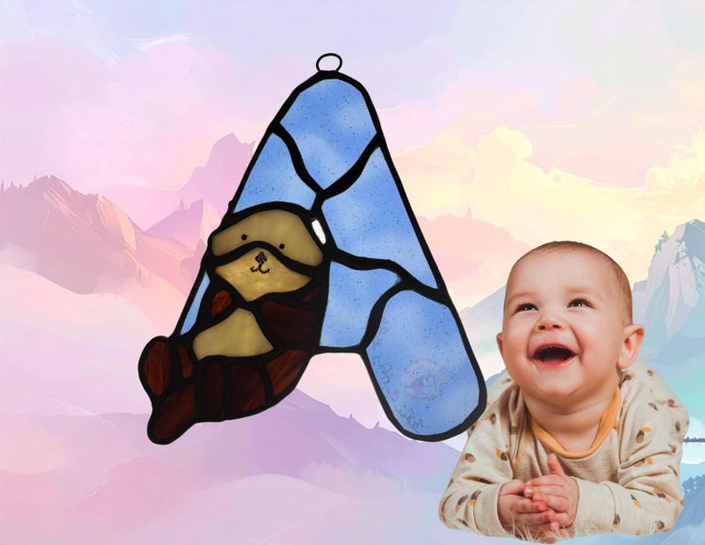 Puede incluir: Un atrapasoles de vidrieras con forma de letra A, con un fondo de cristal texturizado azul claro y una ilustraci&oacute;n de una nutria marina marr&oacute;n y beige. La pieza est&aacute; delineada con soldadura de metal negro e incluye un lazo para colgar.