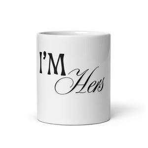 Pode incluir: Uma caneca de cerâmica branca com o texto impresso em preto I'M Hers na parte frontal. A caneca tem um corpo cilíndrico com um acabamento liso e brilhante e uma alça padrão na lateral, oferecendo uma visão clara das letras.