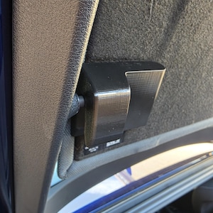 May include: A black, rectangular, 3D-printed plastic bracket mounted to the interior fabric lining of a vehicle. The component features a smooth, layered surface texture and is attached to a small, circular pivot point on the side pillar.
