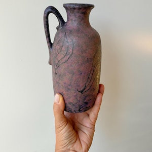 May include: A ceramic pitcher with a mottled purple and black finish, featuring a textured surface and a single handle. The body of the vessel is adorned with subtle, dark, etched leaf-like patterns that wrap around the exterior of the piece.