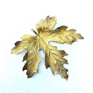 May include: A gold-colored metal leaf with a textured surface. The leaf is shaped like a maple leaf and has a detailed design.