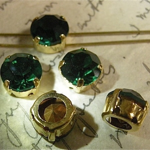 May include: Five emerald green glass rhinestones with gold settings. The rhinestones are round and have a faceted design. The settings are smooth and have a shiny finish.