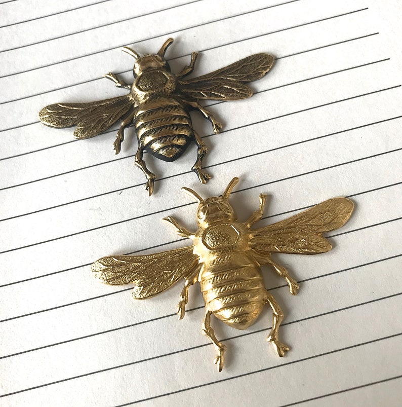 May include: Two gold-colored metal bee brooches. The bee on the left has its wings spread out, while the bee on the right has its wings folded. Both bees have detailed features, including segmented bodies and antennae.